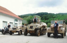 Military vehicles in front of a building with a hill in the background.
