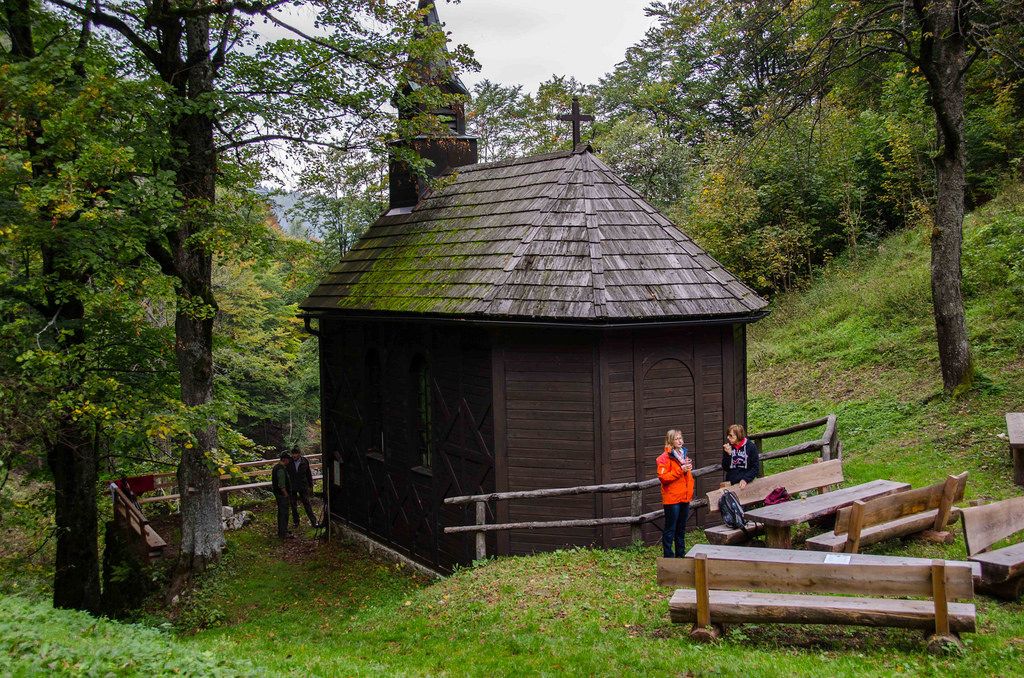 Eine kleine, hölzerne Kirche im Wald mit zwei Personen und Holzbänken im Vordergrund.