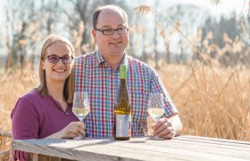Ein Mann und eine Frau sitzen an einem Holztisch im Freien mit Weingläsern und einer Weinflasche.