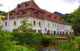 Historisches Gebäude mit rotem Dach an einem Bach, umgeben von grüner Vegetation.