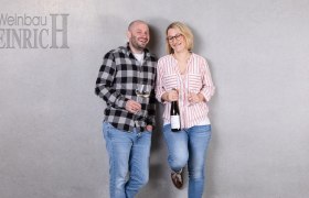 Zwei Personen stehen lächelnd vor einer grauen Wand mit Weingläsern in der Hand. Links steht ein Mann, rechts eine Frau. Oben links steht 'Weinbau Heinrich'.