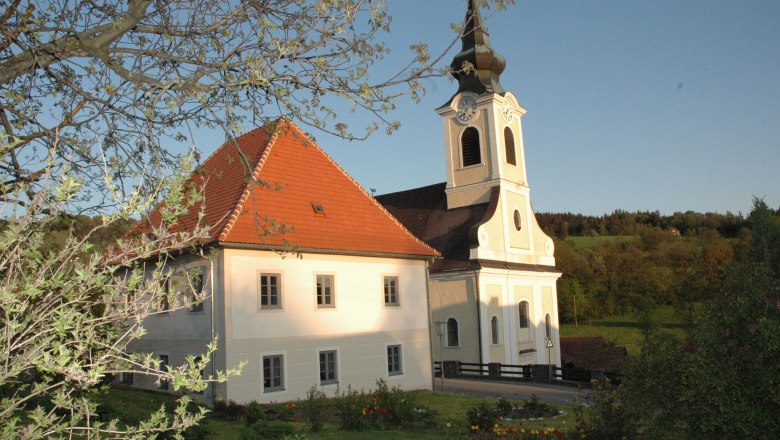Kirche mit Pfarrhof in l&auml;ndlicher Umgebung, umgeben von B&auml;umen und Wiesen.