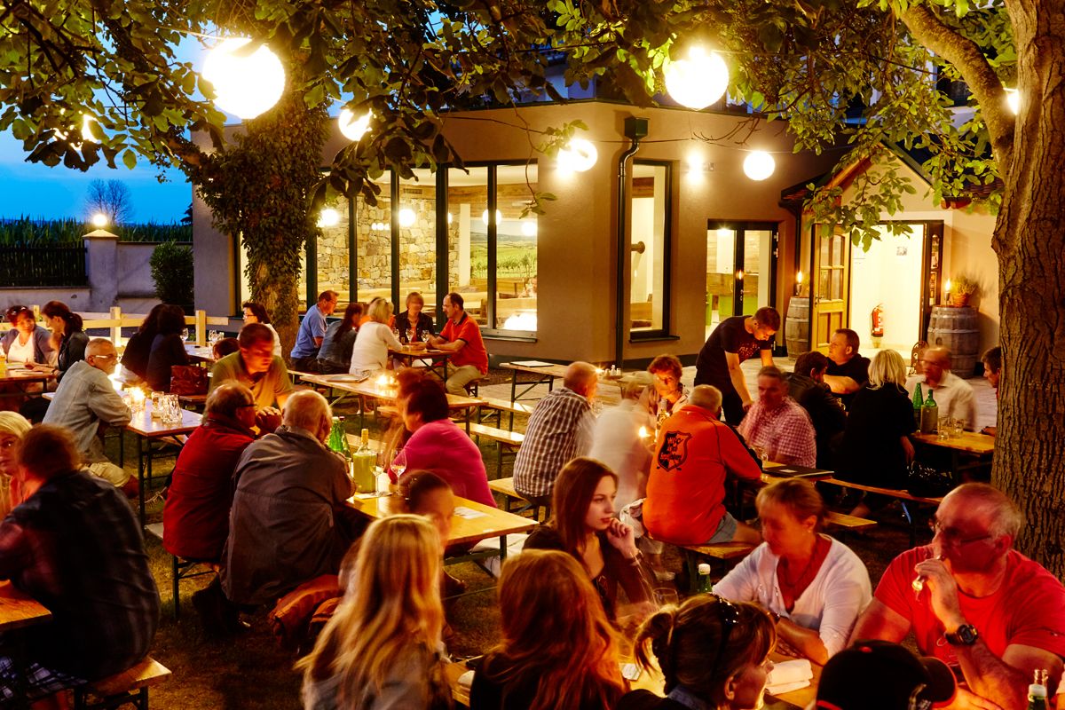 Menschen sitzen abends an Tischen im Freien, umgeben von Bäumen und Lichtern.