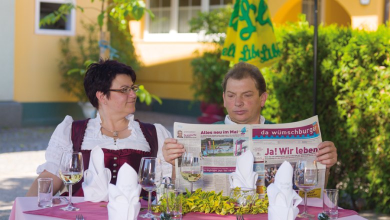 Gasthof Franzl, © Michael Rzepa Ein Mann und eine Frau sitzen an einem Tisch im Freien, der mit Gläsern und Besteck gedeckt ist. Der Mann liest eine Zeitung.