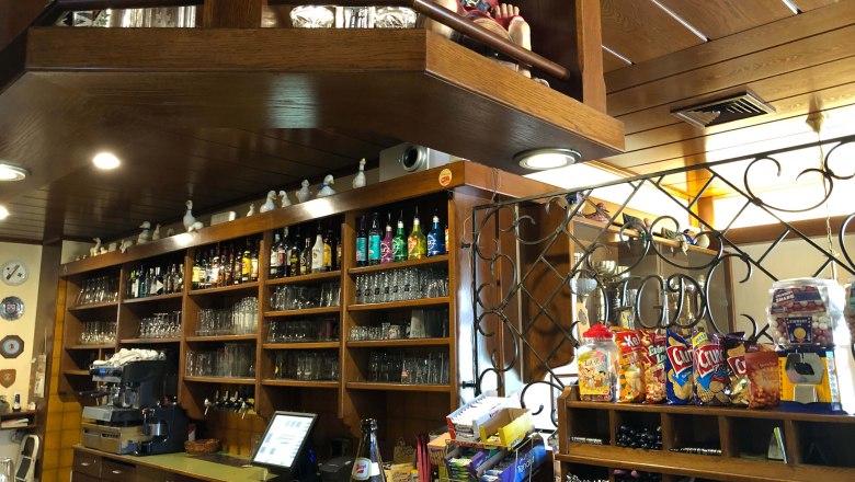 Innenansicht einer Bar mit Holzregalen, Gläsern, Flaschen und Snacks.