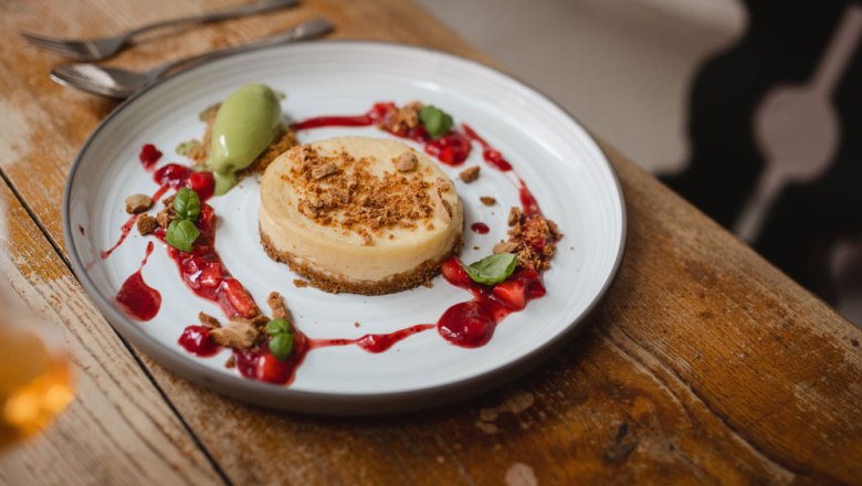 Ein Teller mit einem Cheesecake, dekoriert mit roten Beeren und grünem Eis, auf einem Holztisch.