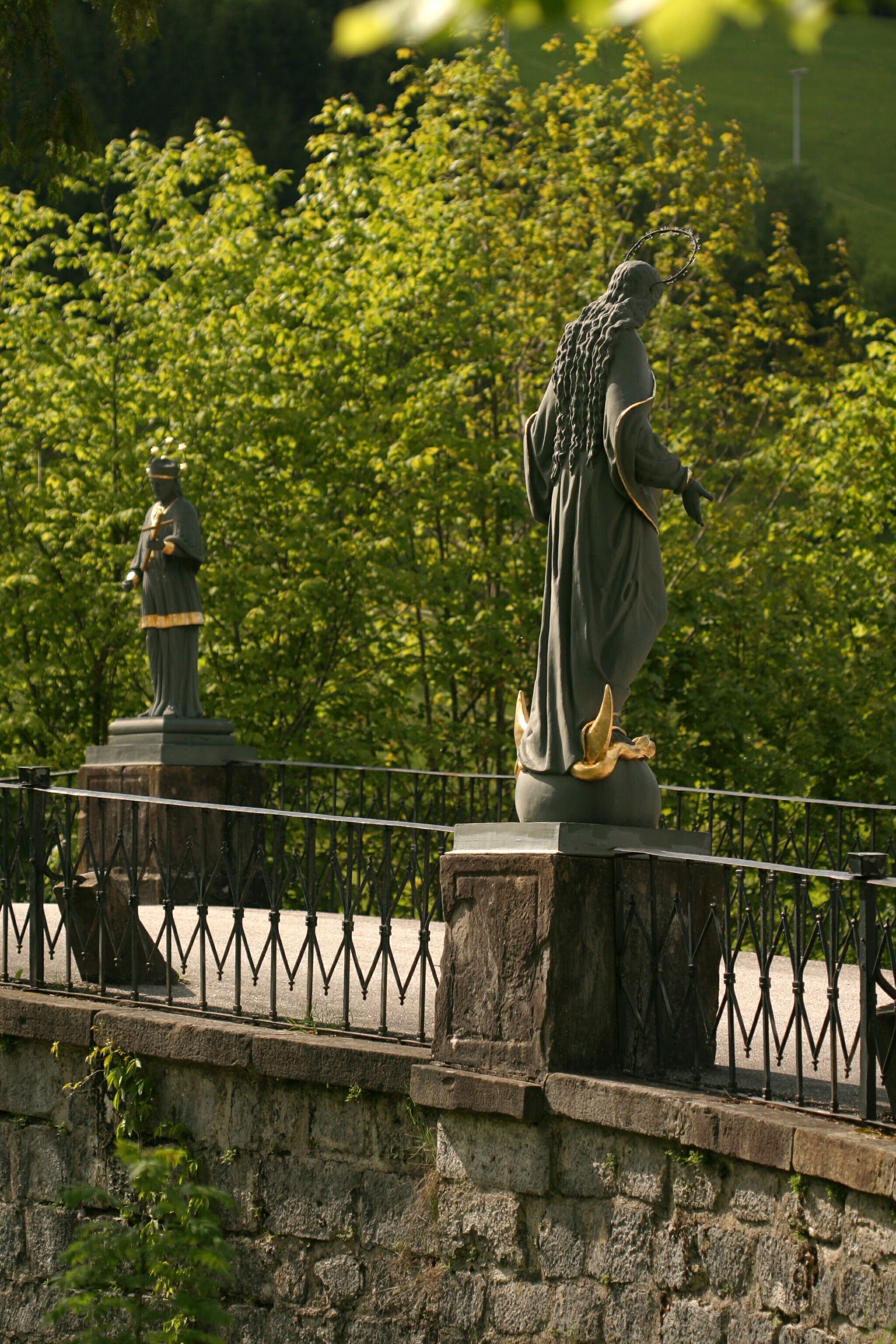 Zwei Statuen auf einer Brücke mit Geländer, umgeben von grünen Bäumen.