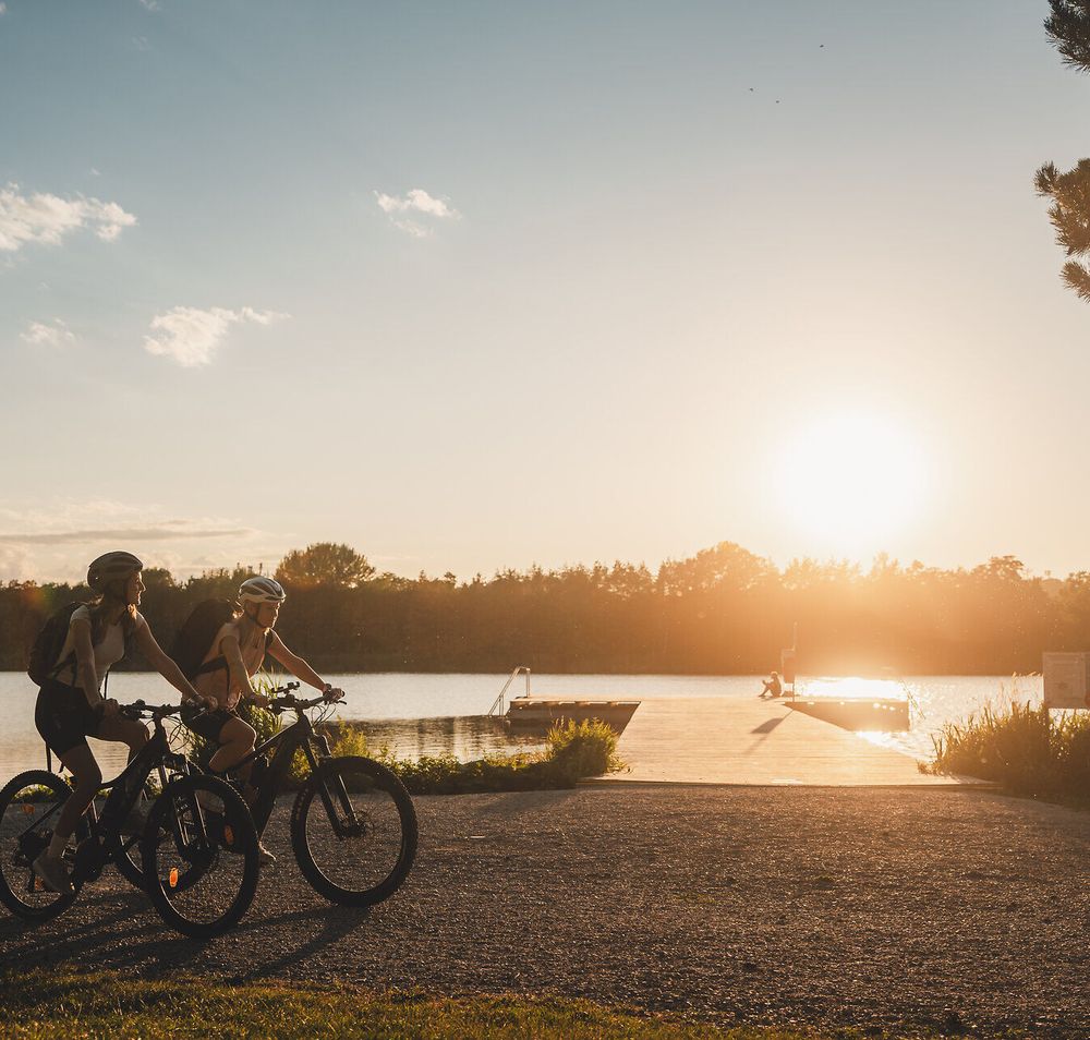 Zwei Radfahrerinnen mit Helmen fahren mit ihren Rädern am Ufer des Viehofner Sees in St. Pölten, im Hintergrund liegt ein Badesteg am Wasser.