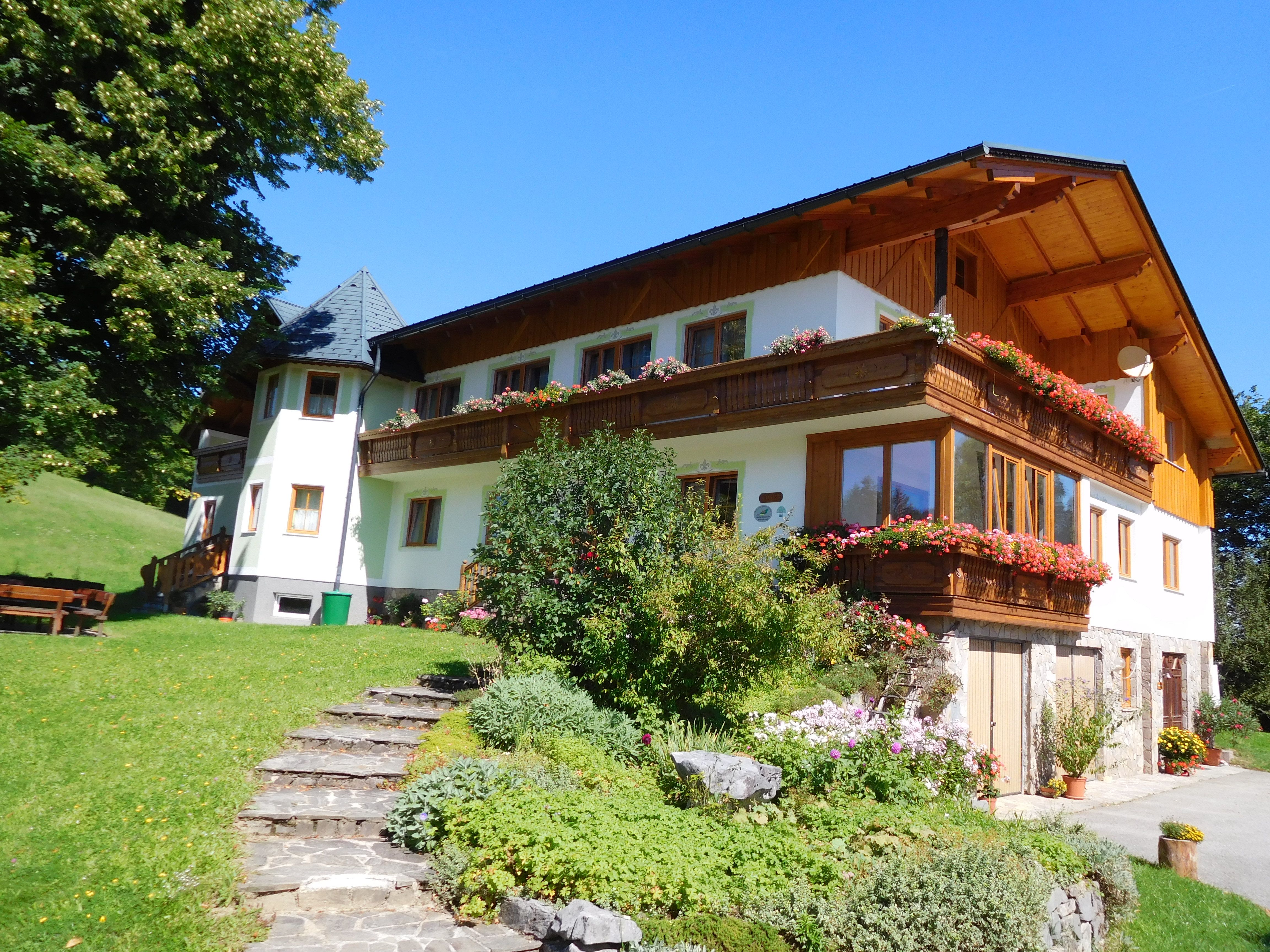 Ein traditionelles Bauernhaus mit Holzbalkon und blühenden Blumen, umgeben von grüner Landschaft und blauem Himmel.