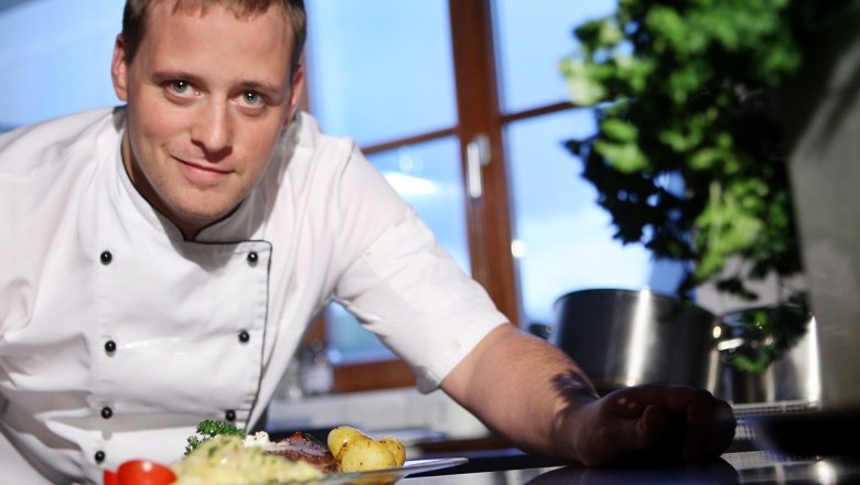 Ein Koch in weißer Uniform präsentiert ein Gericht in einer Küche.