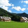 Two wooden houses on a green meadow in front of wooded mountains and a blue sky.
