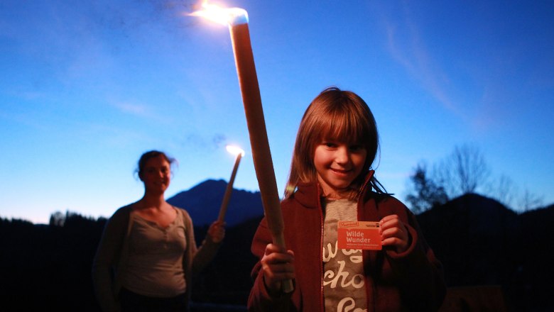 A child holds a burning torch and a map, in the background a woman with a torch, against a mountain backdrop at dusk.