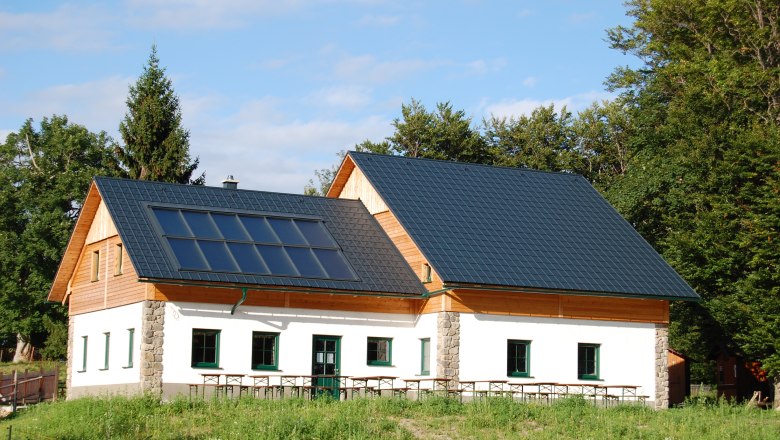 Eine Hütte mit Solarpanelen auf dem Dach, umgeben von Bäumen und Wiese.