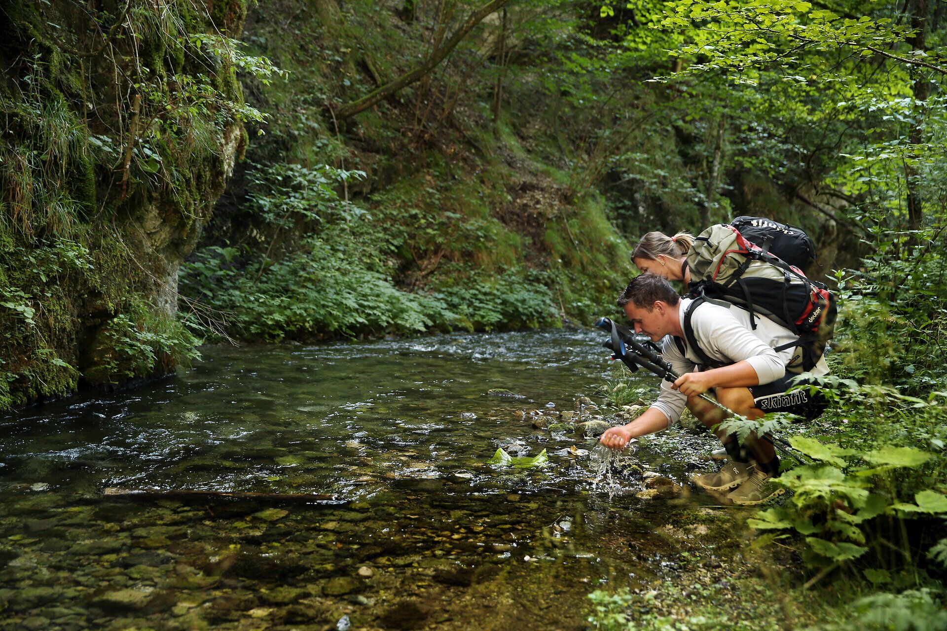 Inmitten der üppigen Natur beugen sich Wanderer über das glitzernde Wasser eines klaren Baches. Umgeben von hohen Bäumen und dem sanften Rauschen des Wassers, spüren sie die Ruhe und Erhabenheit der Umgebung. Diese friedliche Szenerie lädt dazu ein, die Seele baumeln zu lassen und die Schönheit der Natur zu genießen.