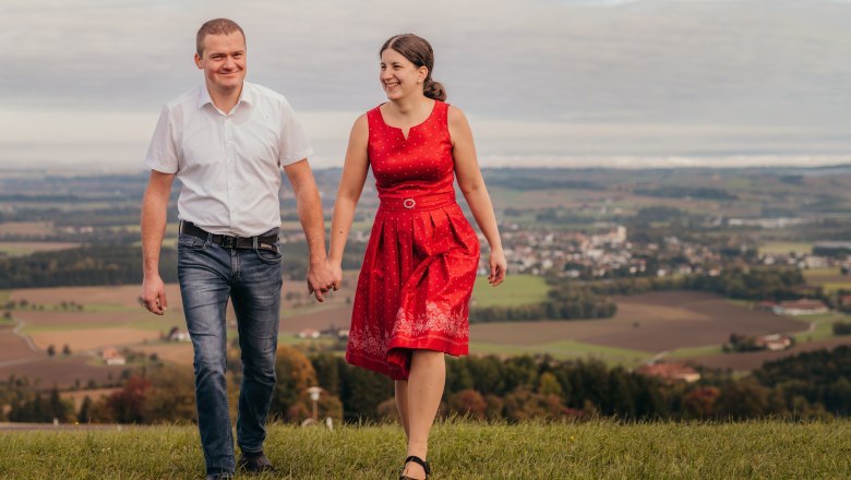 A couple hold hands and walk across a meadow with a landscape in the background.