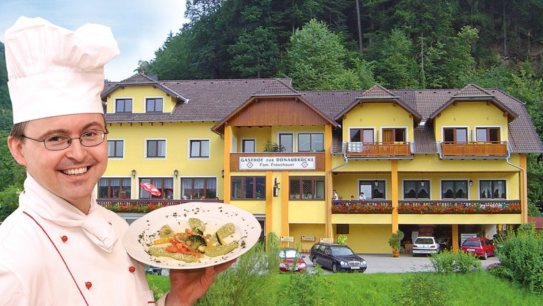 Zur Donaubruecke, © Helmut Froschauer Ein Koch präsentiert ein Gericht vor einem gelben Gasthof namens 'Zur Donaubrücke'.