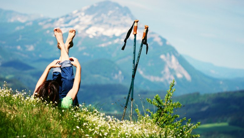 Person liegt auf einer Wiese mit Blick auf die Berge, Wanderstöcke daneben.