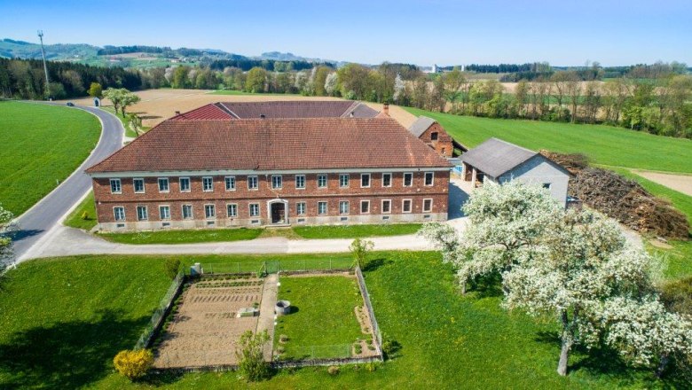 Luftaufnahme eines Bauernhofs mit roten Ziegeld&auml;chern, umgeben von  Feldern und bl&uuml;henden B&auml;umen.