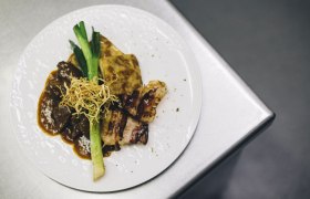 Ein Teller mit Rostbraten, Kartoffelgratin, Lauch und knusprigen Zwiebeln auf einer Metalloberfläche.