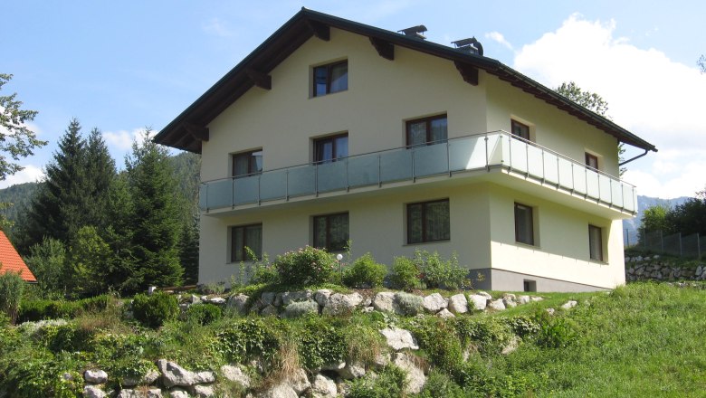 Ein zweist&ouml;ckiges Haus mit Balkon in einer gr&uuml;nen, h&uuml;geligen Landschaft.