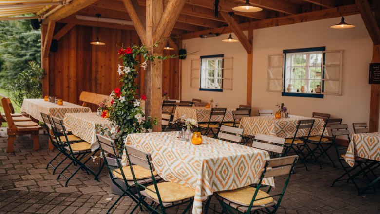 A covered guest garden with wooden tables and chairs, decorated with colorful tablecloths and flowers.