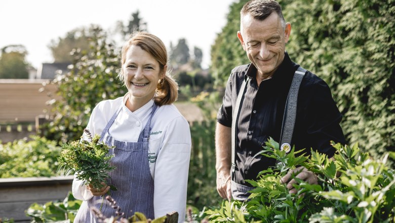 Zwei Personen stehen in einem Garten, eine Frau in Kochkleidung h&auml;lt Kr&auml;uter.