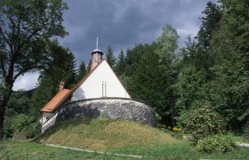 Small church in the forest with a stone base and red roof.