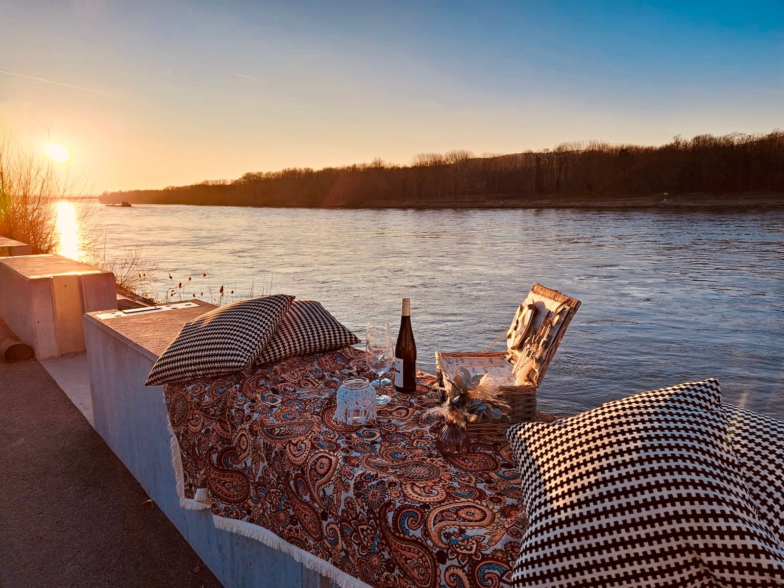 Picknick am Flussufer bei Sonnenuntergang mit Decke, Kissen, Wein und Picknickkorb.