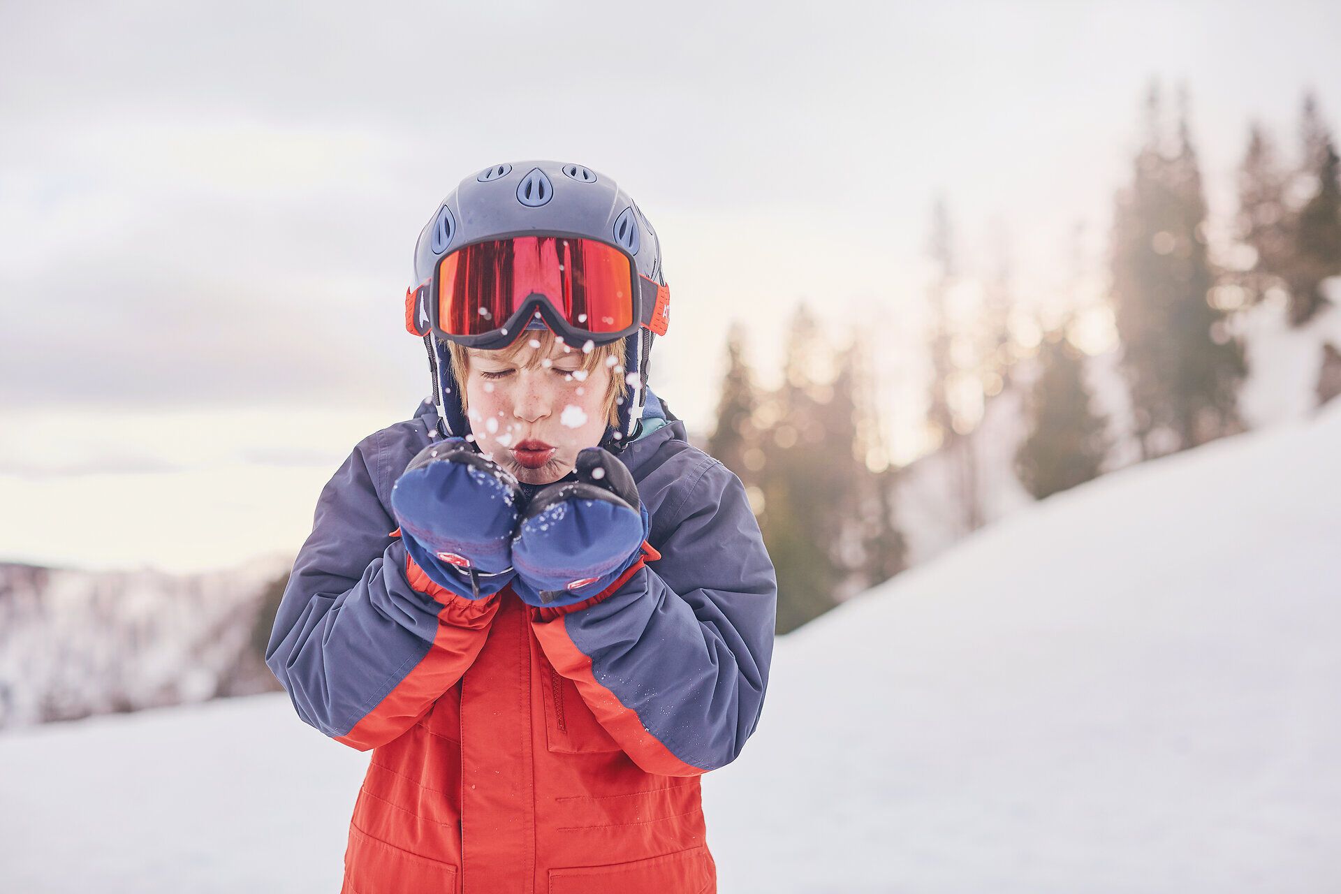 Ein fröhliches Kind genießt den Winterzauber, während es in den schimmernden Schnee bläst. Die strahlende Sonne und die schneebedeckten Berge schaffen eine magische Atmosphäre, die zum Skifahren und Spielen einlädt.