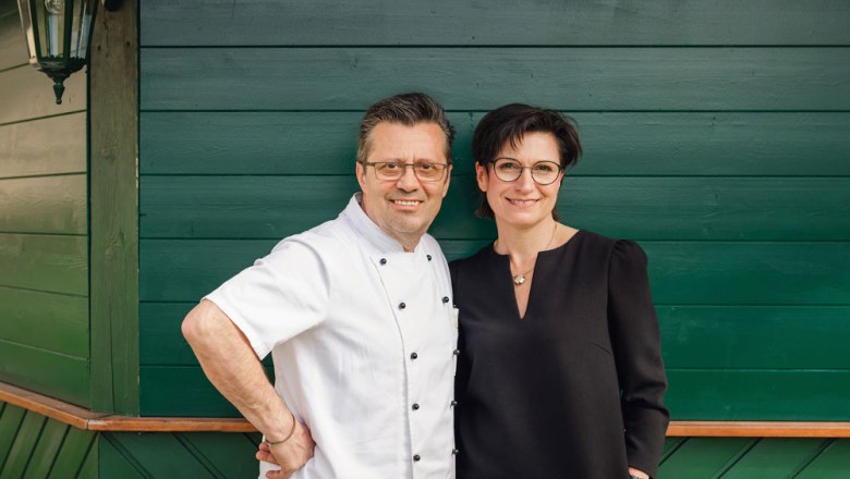 A man in a chef's jacket and a woman in a black dress stand in front of a green wooden wall.