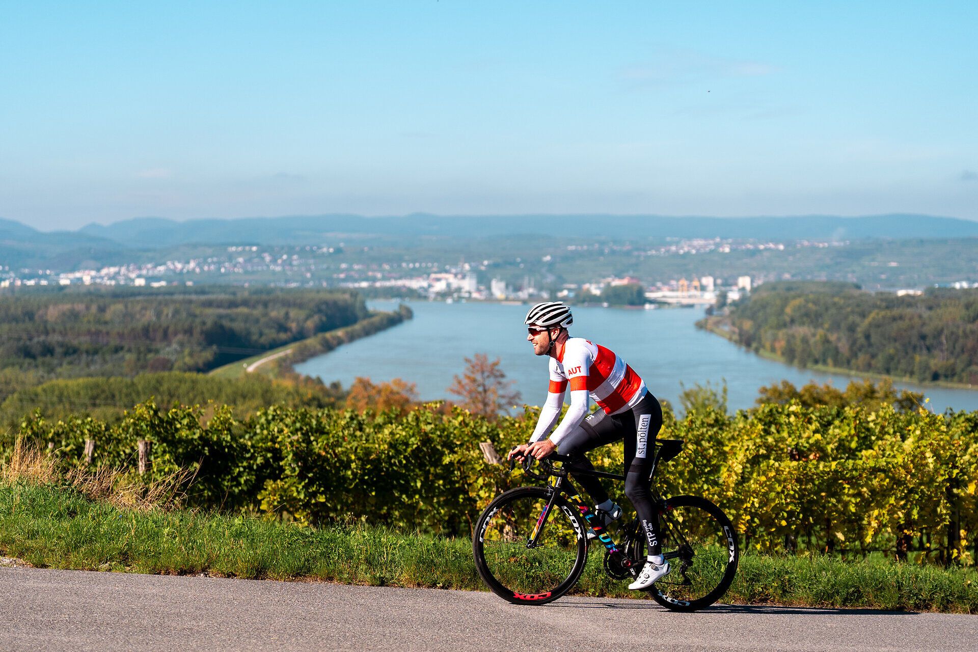Ein Radfahrer genießt die frische Sommerluft und die atemberaubende Aussicht auf die sanften Hügel und den glitzernden Fluss. Die malerische Landschaft lädt dazu ein, die Schönheit der Natur während einer sportlichen Ausfahrt zu erleben.