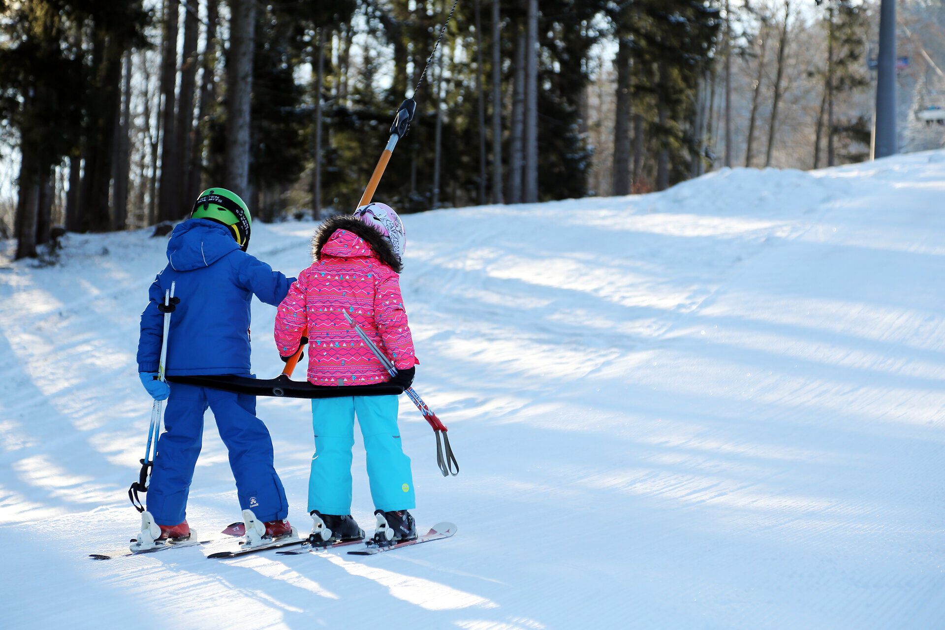 Zwei Kinder stehen auf ihren Skiern, verbunden durch ein Seil, während sie den sanften Hang hinaufgezogen werden. Die strahlende Wintersonne lässt den Schnee glitzern und schafft eine fröhliche Atmosphäre, die die Freude am Skifahren in der Familie widerspiegelt.