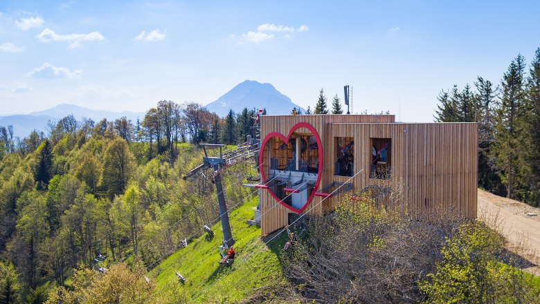Wooden building with heart window and cable car on a wooded hill, mountain in the background.