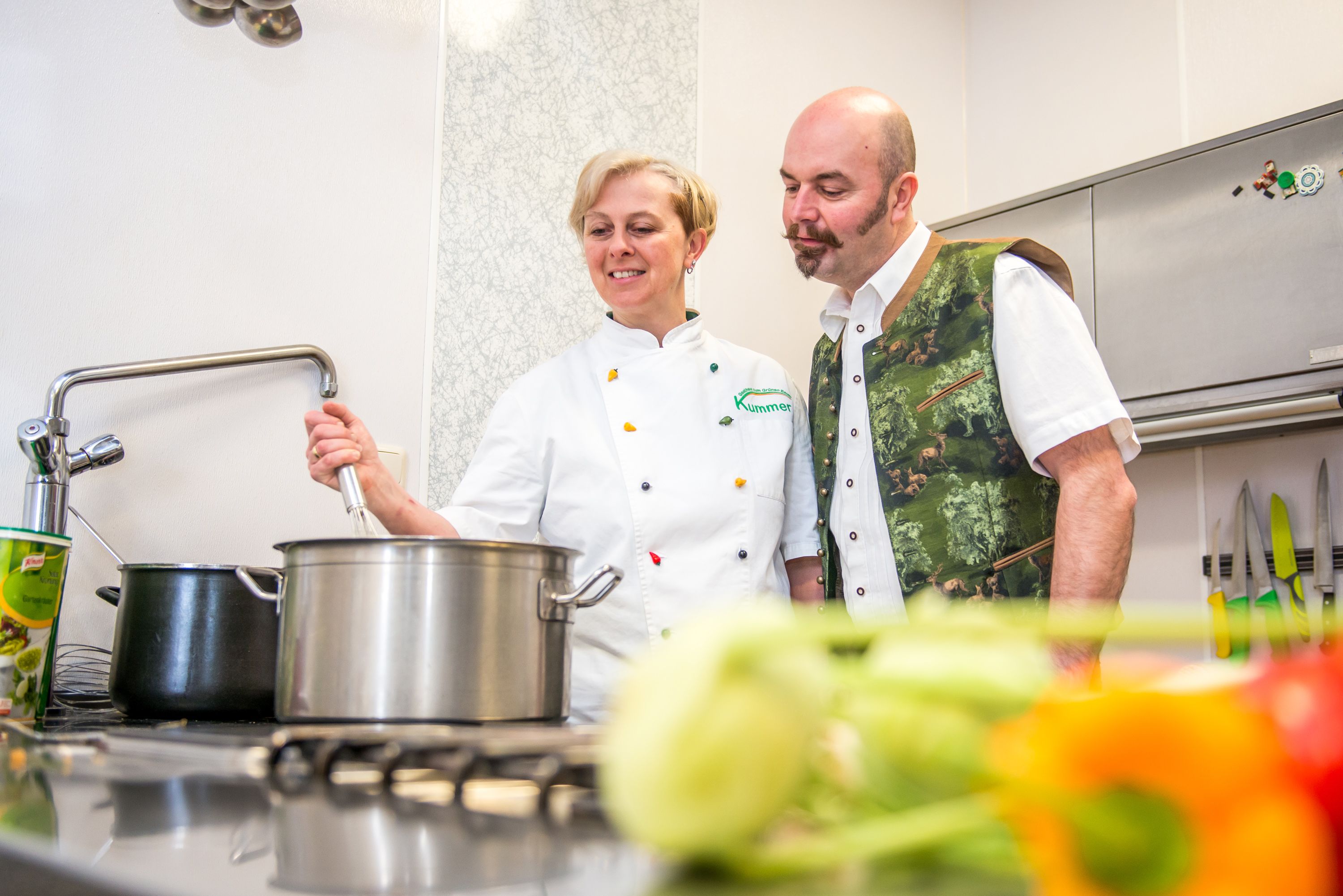 Zwei Personen in einer Küche, eine rührt in einem Topf, die andere schaut zu.