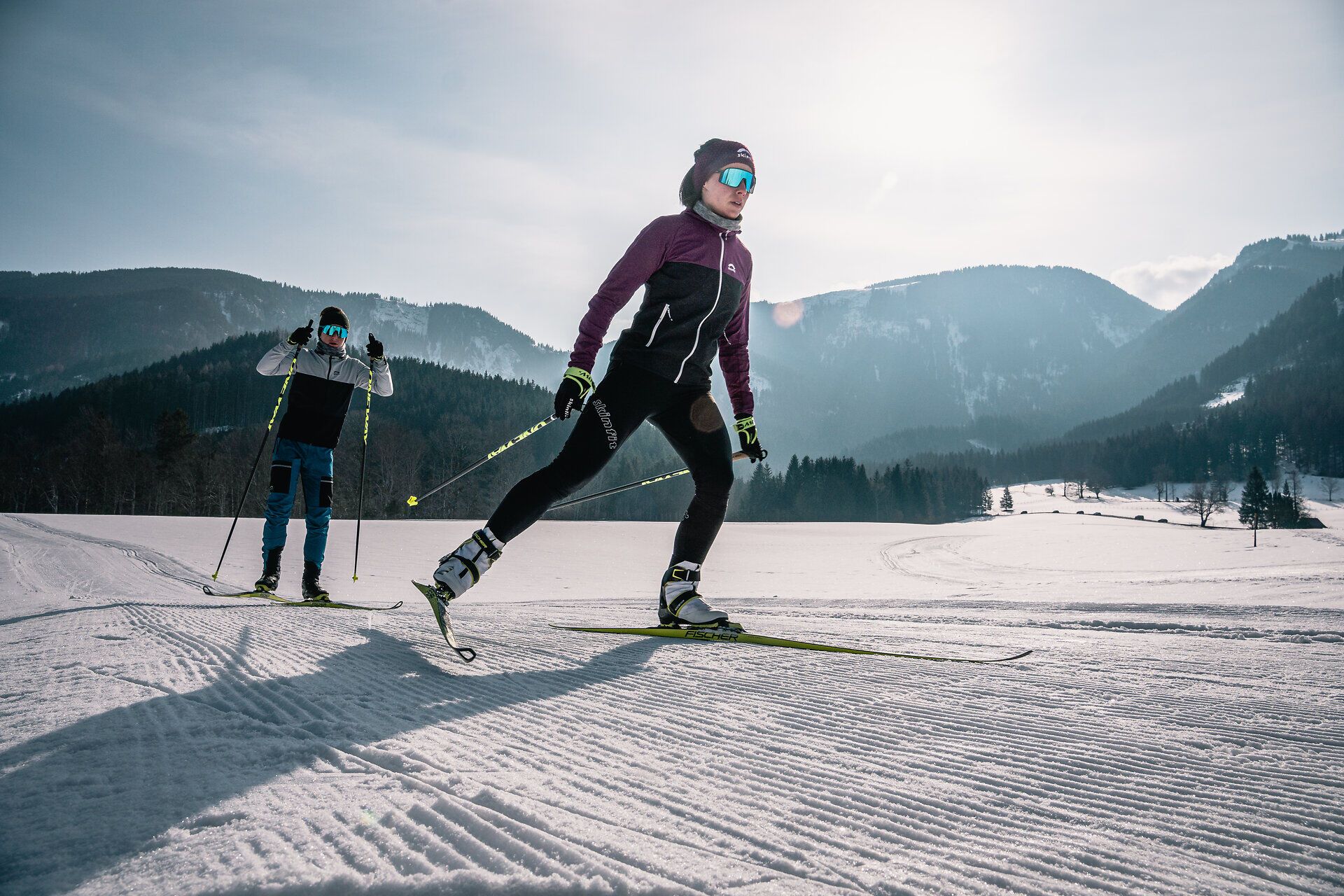 Die glitzernde Schneedecke erstreckt sich über die sanften Hügel der Ybbstaler Alpen, während zwei Skilangläufer in harmonischem Rhythmus durch die winterliche Landschaft gleiten. Die klare, kalte Luft und die majestätischen Berge im Hintergrund schaffen eine perfekte Kulisse für einen aktiven Winterurlaub.