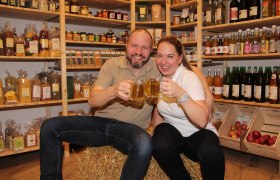 Zwei Personen sitzen auf einem Heuballen im Hofladen und stoßen mit Getränken an.