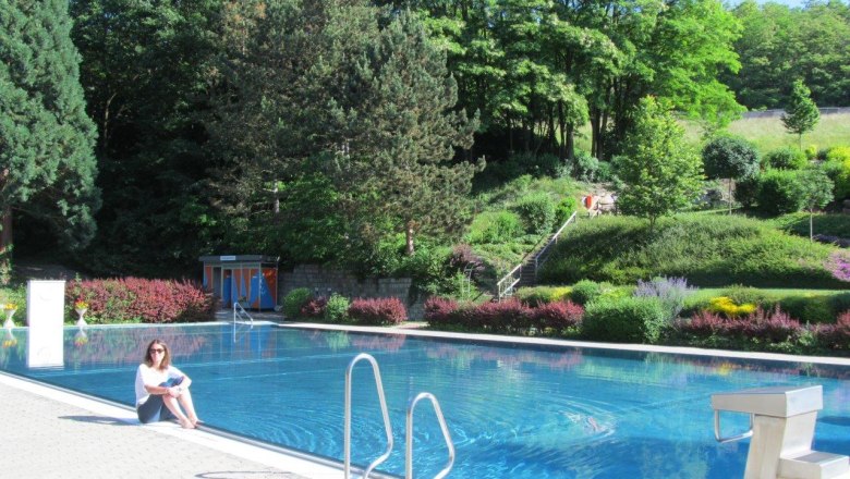 Frau sitzt am Rand eines Freibads mit Wald im Hintergrund.