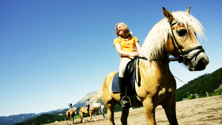 A woman rides a brown horse with a blond mane in a riding arena surrounded by mountains and blue sky.