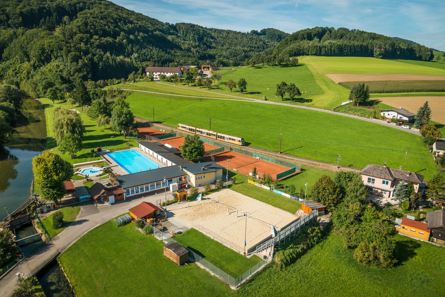 Luftaufnahme eines Freibads mit Pool, Tennisplätzen und Volleyballfeld, umgeben von grünen Feldern und Hügeln.