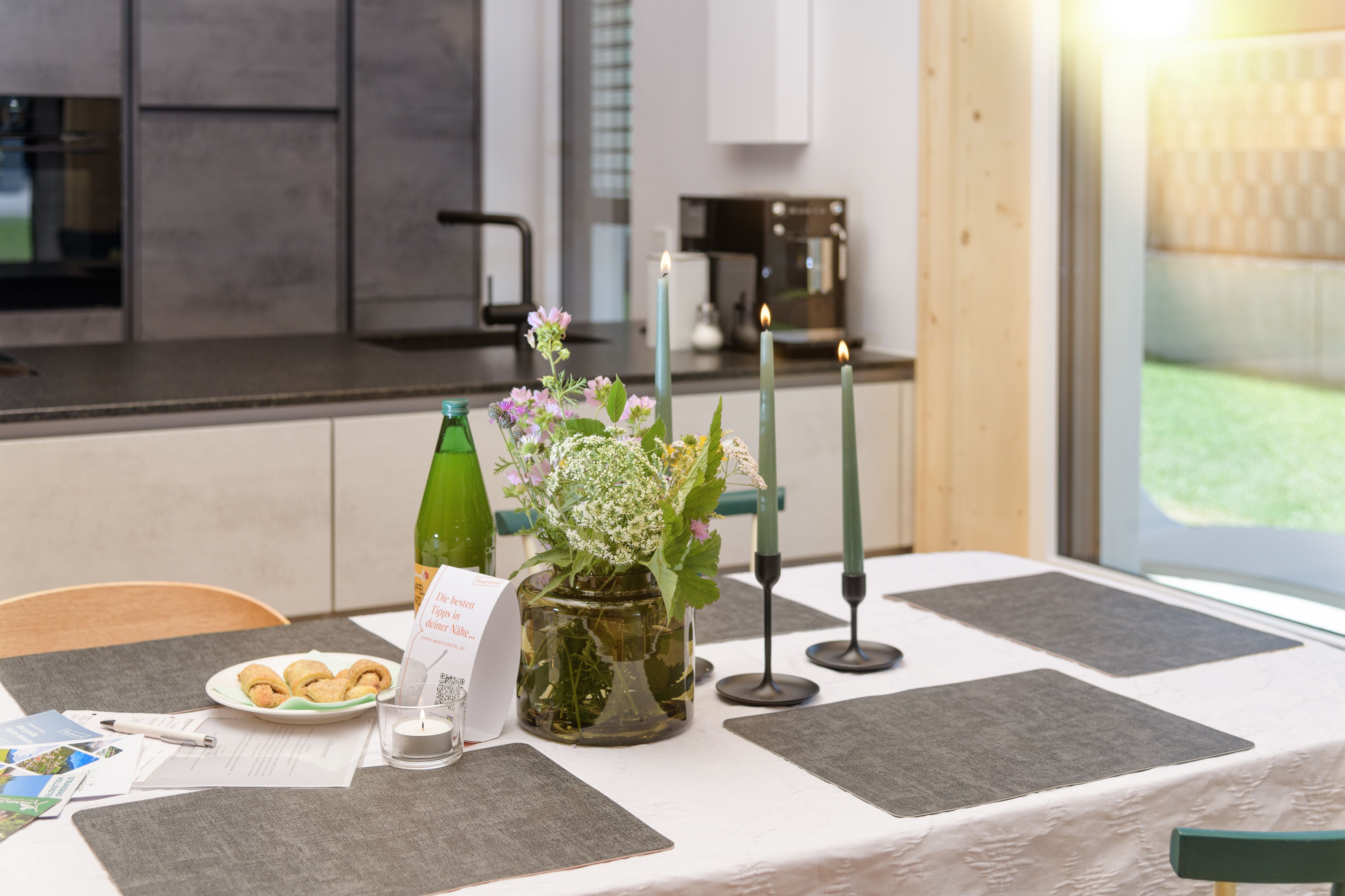 Ein gedeckter Tisch in einer modernen Küche mit Blumen, Kerzen und Snacks.