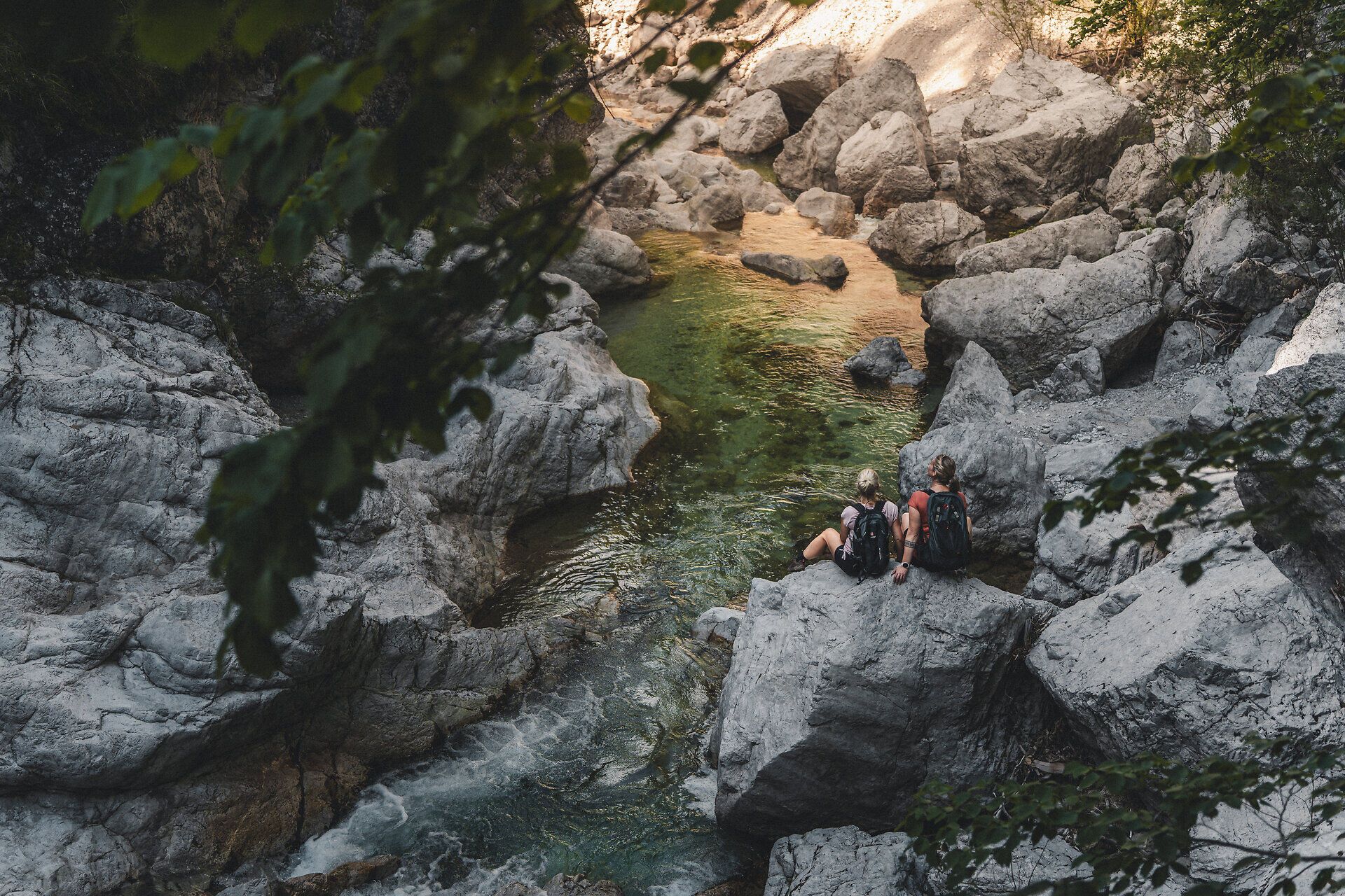 Zwei Frauen sitzen auf großen Felsen an einem klaren Fluss, umgeben von natürlicher Vegetation