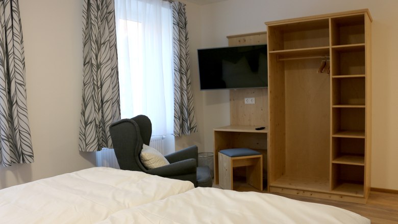 Offener Kleiderschrank mit Fernseher an der Wand. Vor einem Fenster mit einer Gardine steht ein grüner gemütlicher Stuhl.