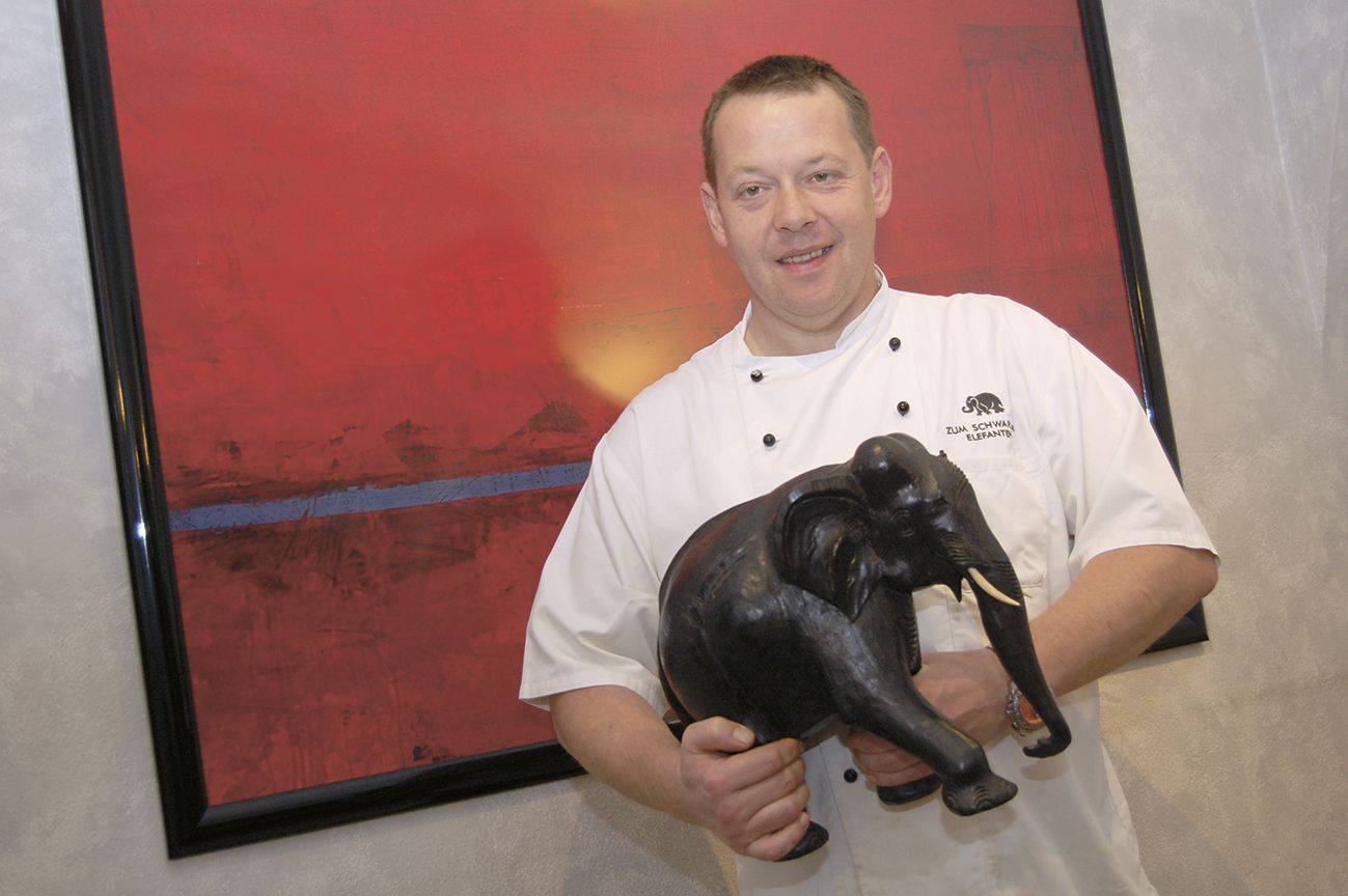 Ein Koch in weißer Uniform hält eine schwarze Elefantenfigur vor einem roten Gemälde.