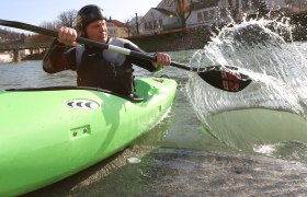 Person in grünem Kajak paddelt auf einem Fluss.
