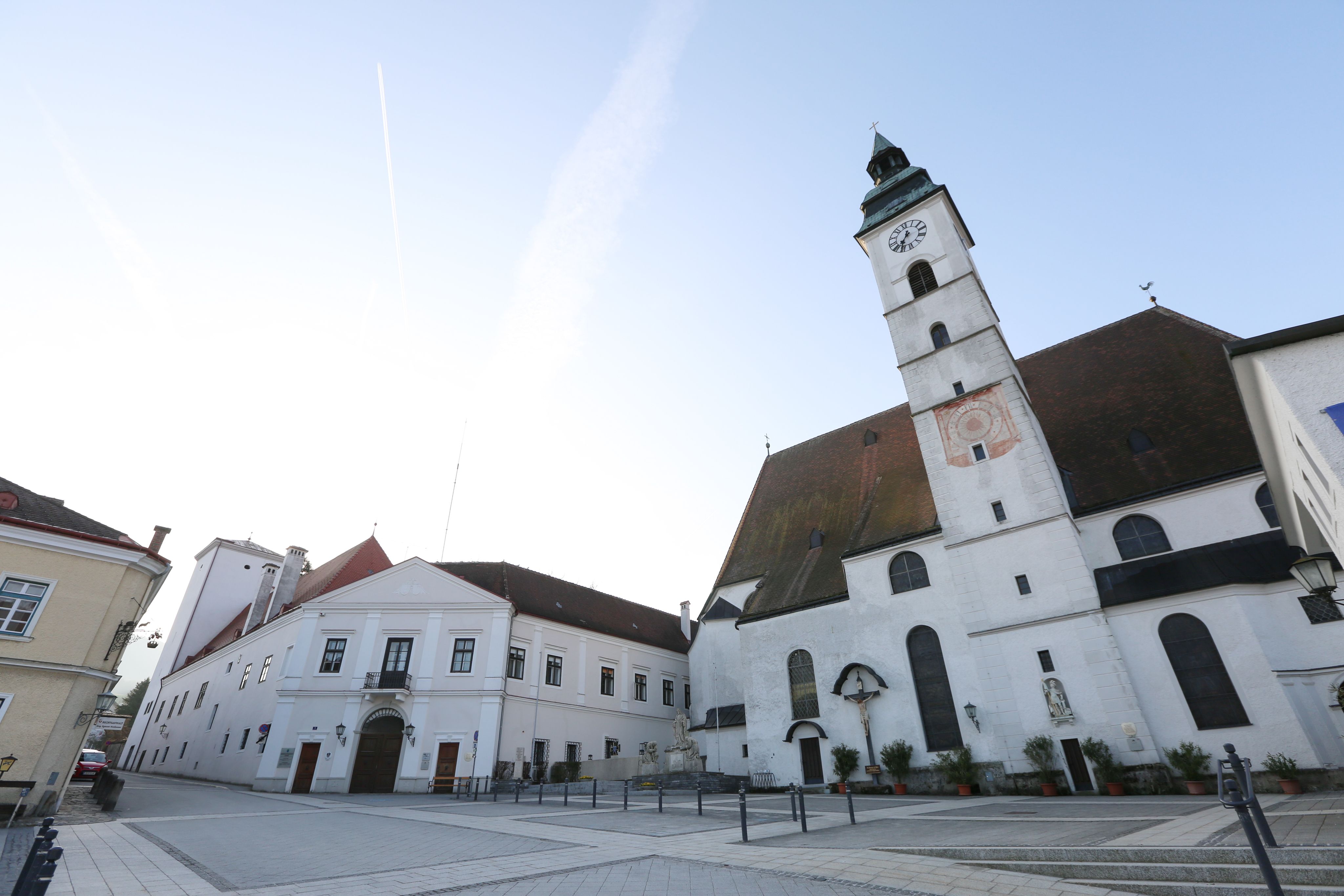Pfarrkirche mit Uhrturm und angrenzenden Gebäuden auf einem leeren Platz.