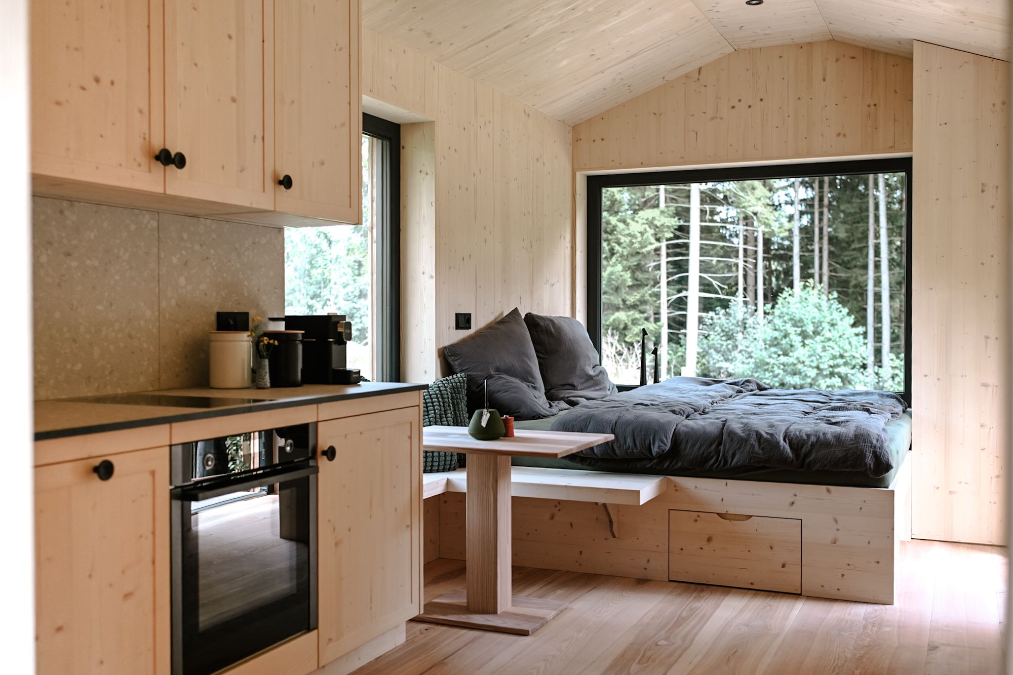 Innenansicht eines modernen Tiny Houses mit Holzverkleidung, einem Bett mit großen Fenstern und Küche.