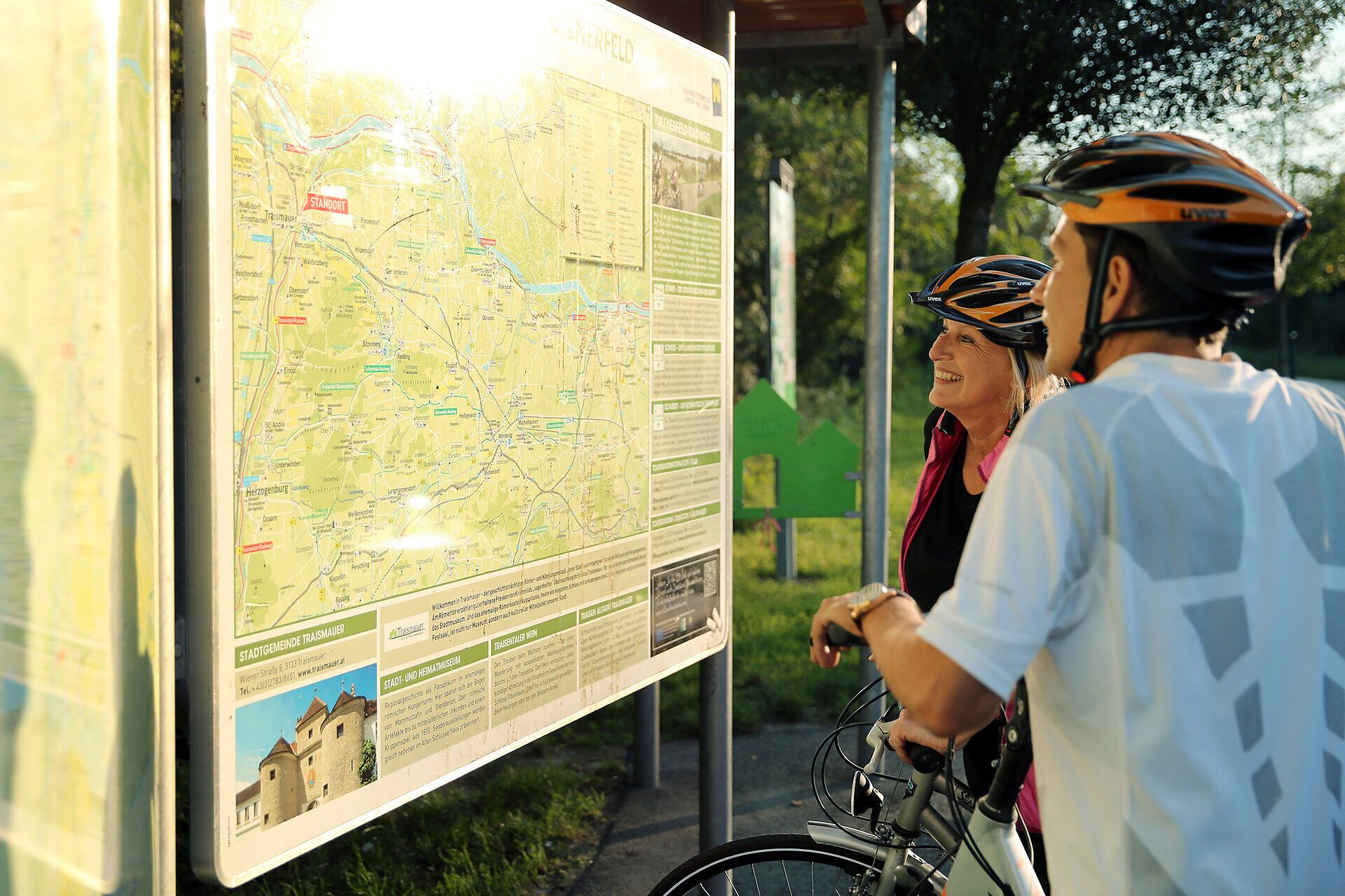 Zwei Radfahrer stehen fasziniert vor einer detaillierten Karte, die die malerischen Routen des Weinbergradwegs zeigt. Umgeben von üppigen Wiesen und sanften Hügeln, spüren sie die Vorfreude auf ein unvergessliches Abenteuer in der Natur. Die goldene Abendsonne taucht die Szene in warmes Licht und verspricht einen perfekten Tag im Freien.