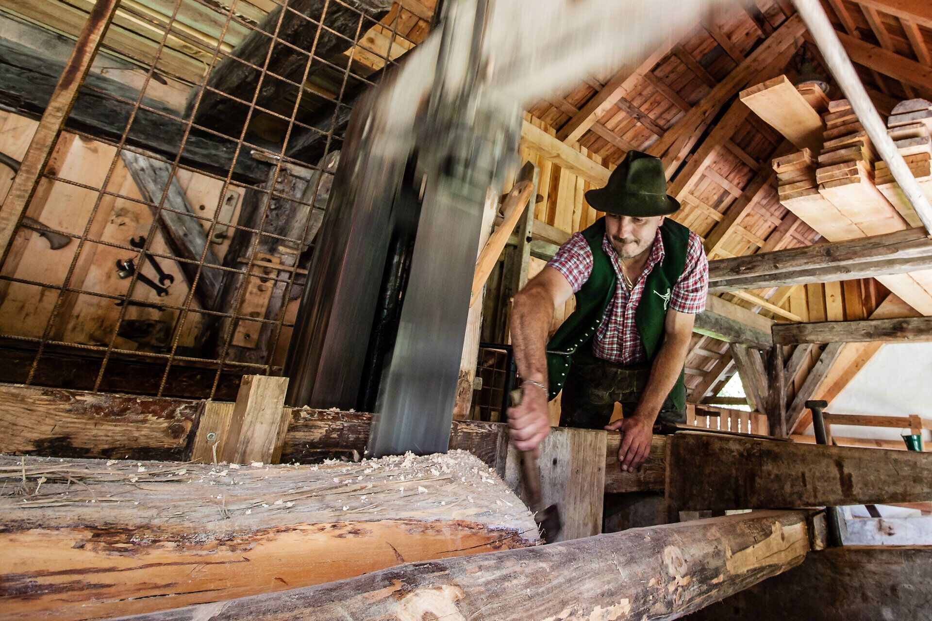 In der Erlebniswelt Mendlingtal wird die traditionelle Holzverarbeitung lebendig. Ein Handwerker arbeitet mit Geschick an der Brettersäge, während die warmen Sonnenstrahlen durch das Dach des alten Holzgebäudes scheinen. Hier spürt man die Verbindung zur Natur und die Geschichte des Handwerks.