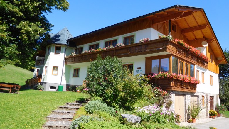 Ein traditionelles Bauernhaus mit Holzbalkon und blühenden Blumen, umgeben von grüner Landschaft und blauem Himmel.