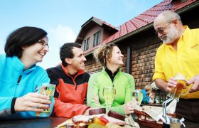 Three people sit laughing at a table in front of a hut while a man in a yellow shirt serves drinks.